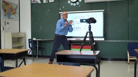 Male teacher using an interactive whiteboard in an empty classroom teaching to a video camera.