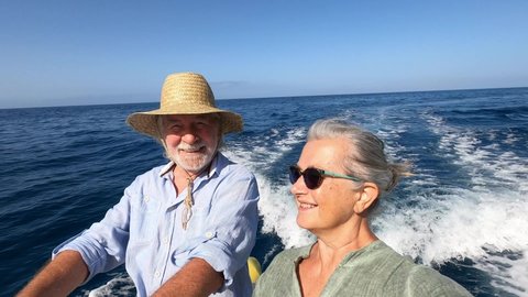 couple of cute mature people or senior enjoying and having fun together in the middle of the sea or ocean with a small boat or dinghy - woman pensioner taking a selfie with her old man driving the boa
