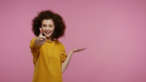 Amazed young woman afro hairstyle in hoodie pointing empty place on her palm, copy space advertising area on hand, expressing excited surprised emotions. indoor studio shot isolated on pink background