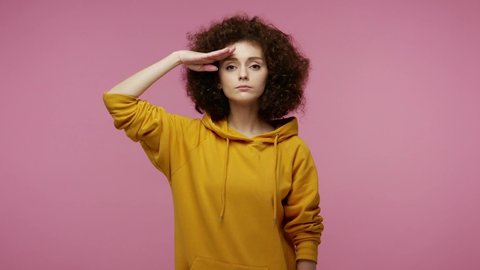 Yes sir! Very responsible patriotic young woman afro hairstyle in hoodie saluting with respect, waiting order from commander, obeying and following discipline. indoor  isolated on pink background