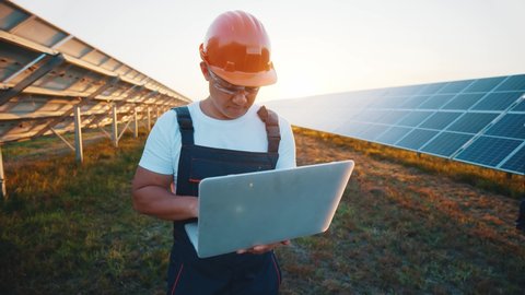 Energy expert asian workman using laptop controlling solar panel batteries supplying electricity on ecological new power generation station. Solar farm concept.