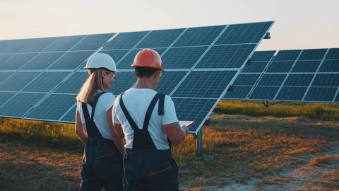 Interracial team of industrial colleagues using tablet monitoring rows of photovoltaic solar panels at sunset. Business cooperation. Solar park. Alternative energy concept.