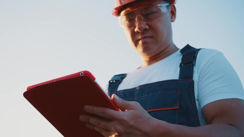 Asian mechanic in uniform wearing hardhat using digital tablet at workspace. Technician calling for help woman engineer discussing plans for construction. Solar farm.