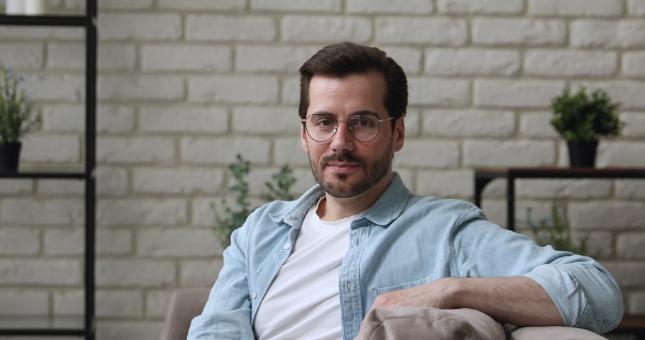 Handsome millennial 30s guy in glasses casual clothes smiling looking at camera sit relaxed on couch at home. Happy homeowner, portrait of attractive man participant of video conference event concept Royalty-Free Stock Footage #1058581222