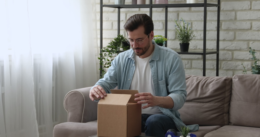 Man sit on couch opens parcel box feels satisfied by delivered goods fragile items. Client buy on-line, e-shopping e-commerce websites services ad, trusted transporting company, quick delivery concept | Shutterstock HD Video #1058581228