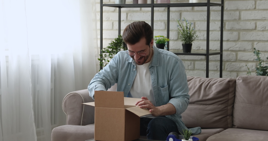Man sit on couch opens parcel box feels satisfied by delivered goods fragile items. Client buy on-line, e-shopping e-commerce websites services ad, trusted transporting company, quick delivery concept | Shutterstock HD Video #1058581228