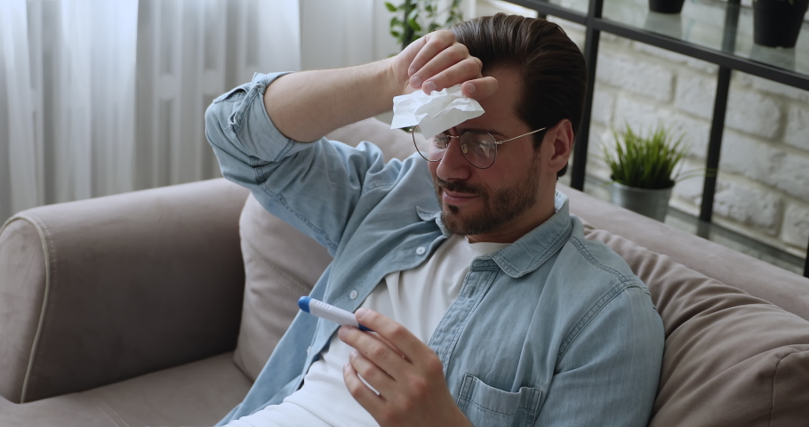 Man sit on sofa wipes runny nose with napkin look at thermometer suffers feels badly due high temperature. Guy catch a cold, influenza virus symptoms, grippe and flue, need care and treatment concept Royalty-Free Stock Footage #1058581264