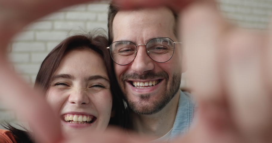 Close up portrait happy couple join fingers together make heart shape with hands smile look at camera through symbol of love standing indoor. St Valentines Day celebration, relations feelings concept Royalty-Free Stock Footage #1058581291