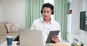 telework concept - Asian man wear headset join a video meeting by laptop and write down information with tablet at home happily