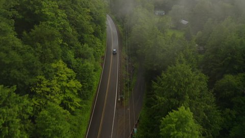 Aerial drone overhead of single car driving down curvy mountain road through green forest valley with trees and rainy fog