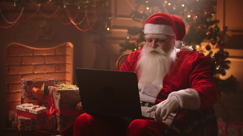 Modern Santa Claus. Cheerful Santa Claus working on laptop and smiling while sitting at his chair with fireplace and Christmas Tree in the background. | Shutterstock HD Video #1058591650
