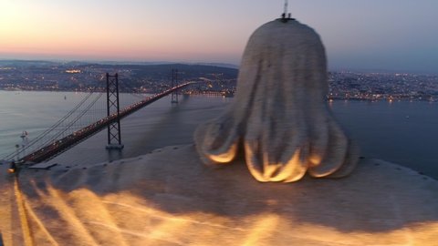 Aerial view tremendous monument of Christ in twilight. Drone flyby pass near crop stone head illuminate in twilight and overlooking Tagus river and 25th april bridge with driving cars.