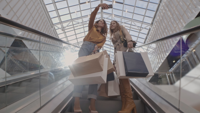 Low angle view of two young female friends with several shopping bags in their hands standing on escalator and making selfie using smartphone camera Royalty-Free Stock Footage #1058596579