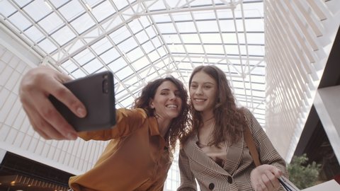 Two young Caucasian women standing in shopping mall, holding several shopping bags in their hands and making selfie using cellphone camera