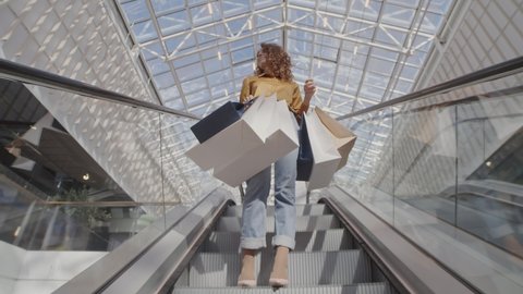 Low angle view of young Caucasian woman wearing trendy clothes standing on escalator with several shopping bags in her hands