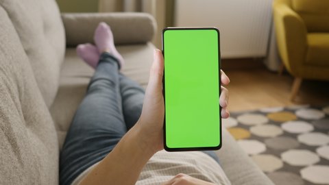 Young Woman Home Lying on a Couch with Green Screen Smartphone in Vertical Mode. Girl Using Touch Green Screen Mobile Phone. Female Using Smartphone, Browsing Internet, Watching Video Content, Blogs. 