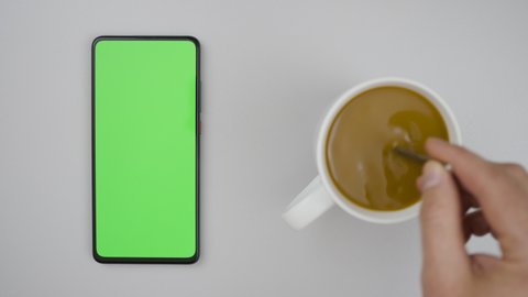 Man Hand Cup of Hot Coffee and Using Smartphone Watching Green Screen Top View. Smartphone with Green Mock-up Screen Business Concept. Person Hand Stirring Coffee with Spoon on Table.