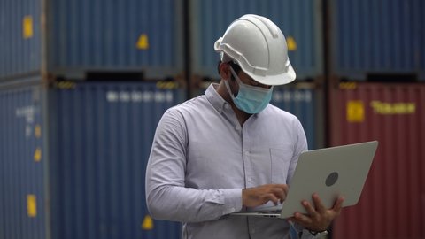 Foreman wareing face mask holding laptop computer talking on the walkie-talkie control loading Containers box at warehouse logistic in Cargo . during coronavirus pandemic or covid 19 outbreak 
