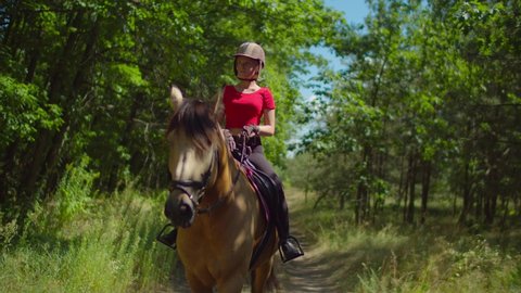 Serious attractive teenage girl jockey in helmet and horse riding clothing enjoying horseback ride on brown purebred horse on dusty trail in summer nature, expressing confidence and determination.