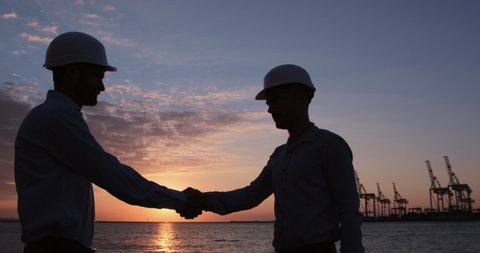 Engineers discuss the work plan on the tablet and shake hands while standing in the seaport against the backdrop of the sunrise. Agreement to do work between people.