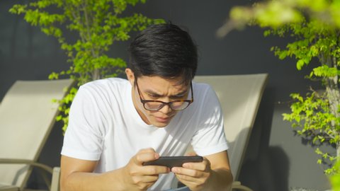 close up teenager asian man sitting at outside home and holding smartphone to play game tournament final round online and feeling excited after become a winner for lifestyle technology concept
