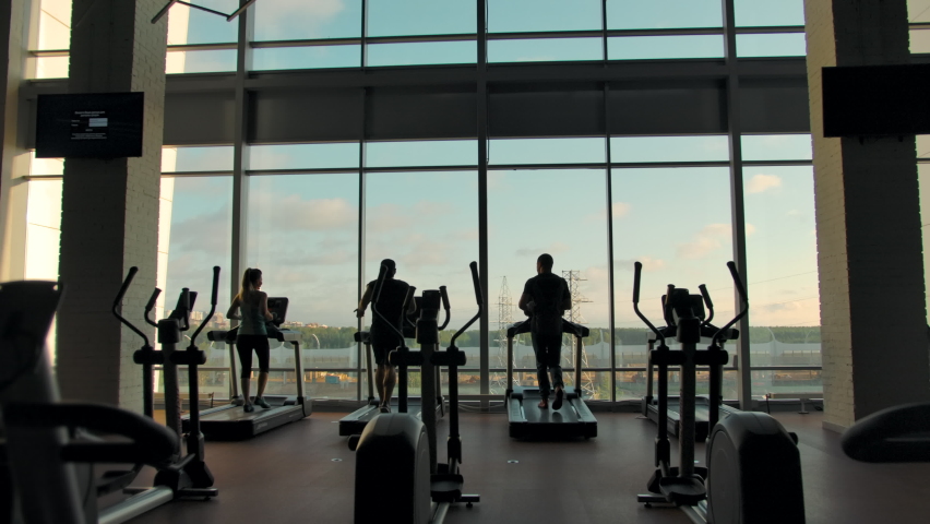 Three unrecognizable people running on treadmills in an empty gym against a large window on a sunny day. Right to left smooth slow motion dolly shot. | Shutterstock HD Video #1058604208