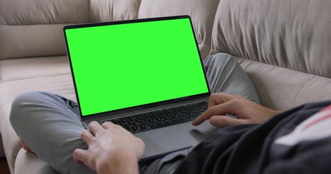 Man Uses MacBook Pro 16 inch Laptop with Green Mock-up Screen While Sitting at the Sofa in His Cozy Living Room. Pre Keyed Chroma Key 