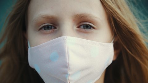 Portrait of little girl looks upset at camera, child wears protective medical mask on his face against infection covid virus, small lady stands on street during pandemic epidemic of disease, close up