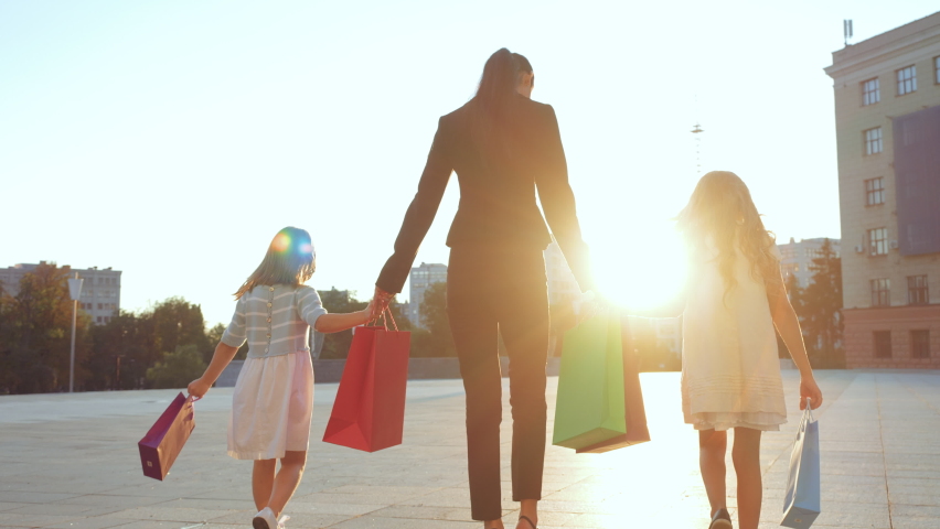 Young mom brunette wife walks in stylish business black suit, holds hands of two daughters girlfriend in new white baby dresses bought in store, carries bright shopping bags, family shoppers rear view Royalty-Free Stock Footage #1058604577