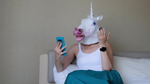 Woman with a head mask of unicorn talking to web camera on smart phone on a bedroom. Social media concept. Weird funny video