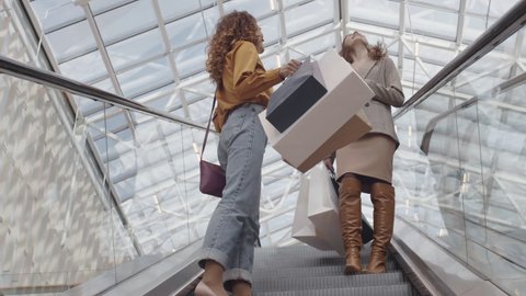Low angle view of two joyful young women with shopping bags and drinks in their hands going down on escalator in store while talking