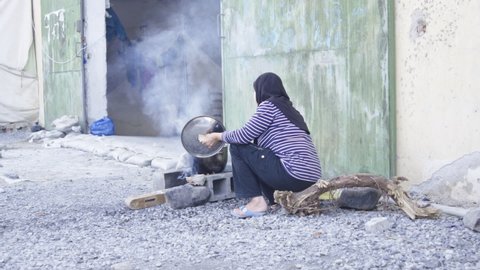 A Syrian woman cooks in a pot on top of stones arranged as an improvised camp fire, while sitting in a trunk tree. Refugee camp in Greece