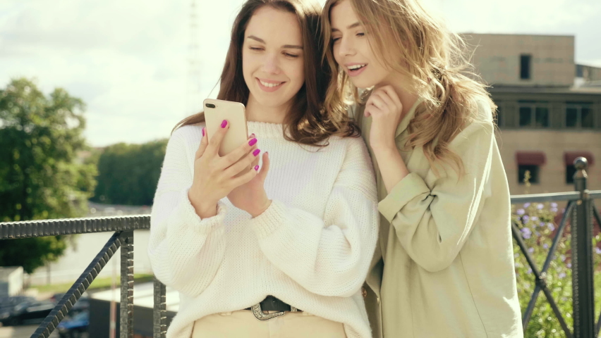 Two young beautiful smiling hipster girls.Sexy carefree women posing near wall in the street in hats. Positive models having fun.They looking at cellphone screen laughing enjoying using mobile apps