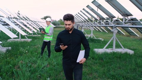 Businessman while perge talking on the phone near the construction of solar batteries. He fell out of motion. Happy young man discussing on the phone the opportunity to expand the solar panels.