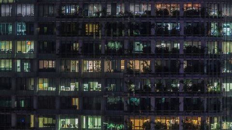 Modern office building with big windows at night timelapse close up view, in windows glowing light shines and some people inside, Singapore. Zoom out