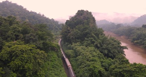 Aerial view of Passenger train pass through the middle between mountains among the forest parallel the river with scenic landscape in green season have foggy at Kaeng Luang, Phrae, amazing Thailand