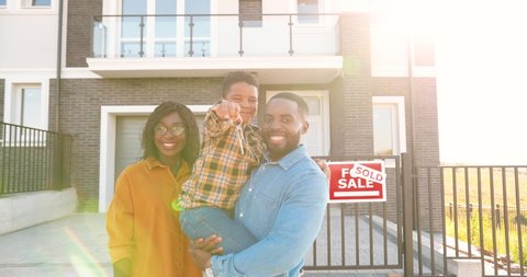 Portrait of happy African American family with small child standing at new house at suburb and demonstrating keys to camera. Cheerful parents and son showing key and moving in new home. Sold.