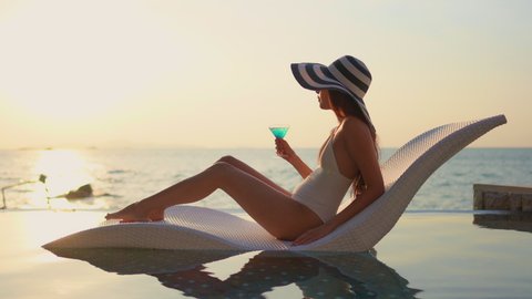 Pretty woman in white bathing suit and wide hat reclining in modern chair in swimming pool overlooking a beach at sunset. Lady with colorful tropical drink crosses legs watching ocean.