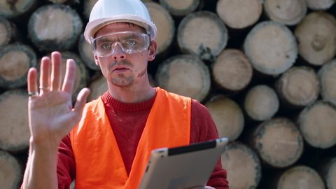 An engineer wearing safety goggles uses tablet computer harvest logs. Communication working personnel when unloading tree logs at plant. Coordination work in forest for timber loading. 