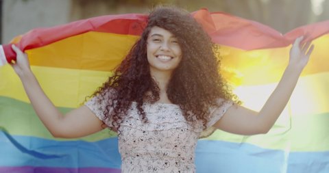Young curly hair woman covering with lgbt pride flag. Alone. One. Keeping fist up, covering LGBT flag. LGBT+ flag on outdoor background. 4K.