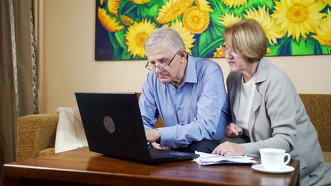 Serious pensioner aged husband and wife manage utility bills using laptop at home, concerned senior couple read bank loan or mortgage documents at table, elder man and woman check insurance paper