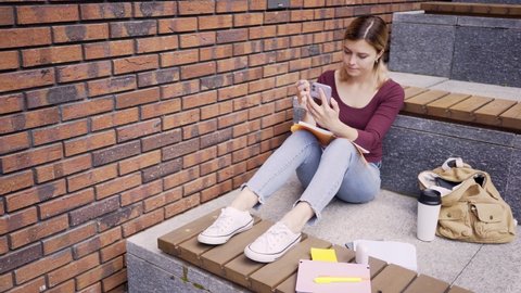 young stressed woman student scores out something in paper notebook with pen looking at smartphone screen on stone bench on modern city street