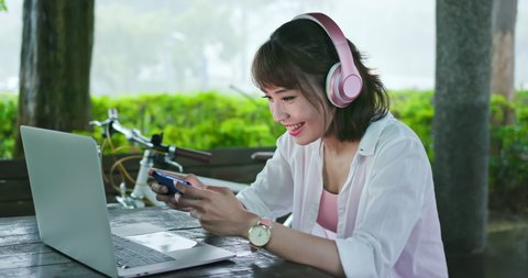 Asian woman student wear headphone play mobile game in the park with laptop in the park happily