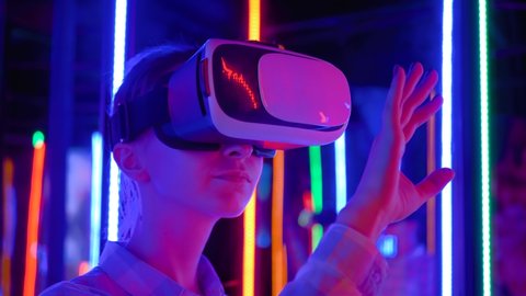 VR, futuristic, retrowave, immersive, entertainment concept. Slow motion: woman using virtual reality headset and looking around at interactive technology exhibition with colorful illumination