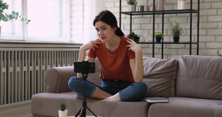 Young woman sitting cross-legged on couch at home put smart phone on tripod talking by video call. Counselling on-line psychological counselor provide help to client distantly, record new vlog concept Royalty-Free Stock Footage #1058642092