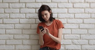 Smiling girl in casual clothes stands against white brick wall background use cellphone chatting with boyfriend online, enjoy shopping on internet. Modern tech and young generation, websurfing concept