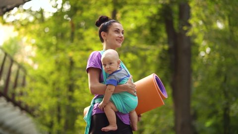 Young woman carrying baby in sling while walking in green park, yoga mat under her arm. Athletic mother going to outdoor training with little child. Concept of fitness