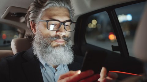 Handsome middle aged businessman with grey hair and beard riding in the backseat of taxi cab, surfing the Net on smartphone and looking at city street through the window