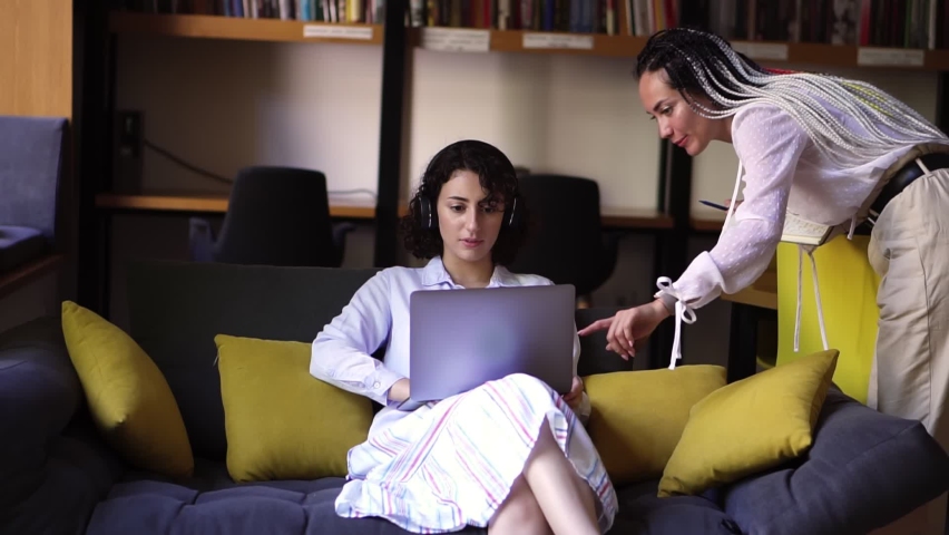 Businesswoman working on laptop at home office. Young female professional working on computer device. Bookshelves with books in the background. Sitting on sofa with headphones, her friend showing smth Royalty-Free Stock Footage #1058645944