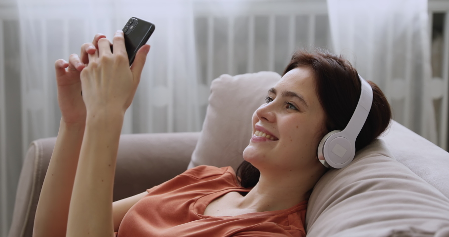 Close up women wear headphones hold smartphone lean on couch in living room relaxing on lazy weekend enjoy distant chat with friend listen to music. Hobby and leisure using modern wireless tech | Shutterstock HD Video #1058647837